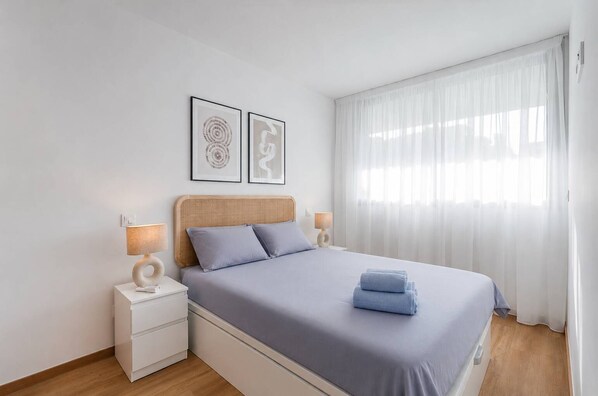 Een slaapkamer met een bed, twee nachtkastjes, een lamp, een raam met gordijnen en twee ingelijste foto's aan de muur.