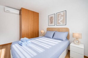Een hotelkamer met een bed, een nachtkastje, een wandairconditioner en twee ingelijste kunstwerken.