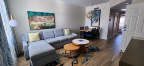 Un salon comprenant un canapé d’angle, une table à manger avec des chaises, une table basse ronde, un tableau au mur et un tapis à motifs.