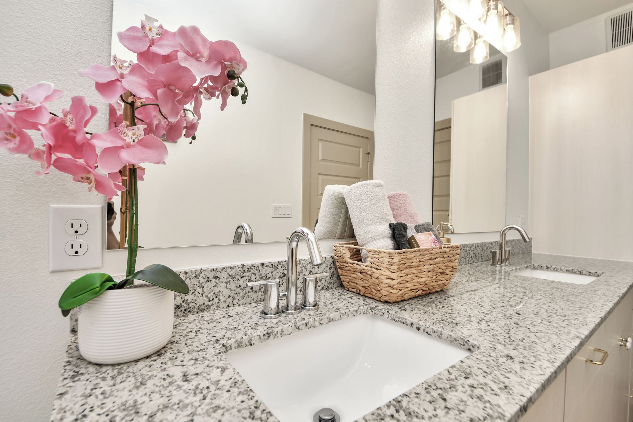 Une salle de bain avec un comptoir en granit, deux lavabos, un miroir, une orchidée en pot et un panier rempli de serviettes.