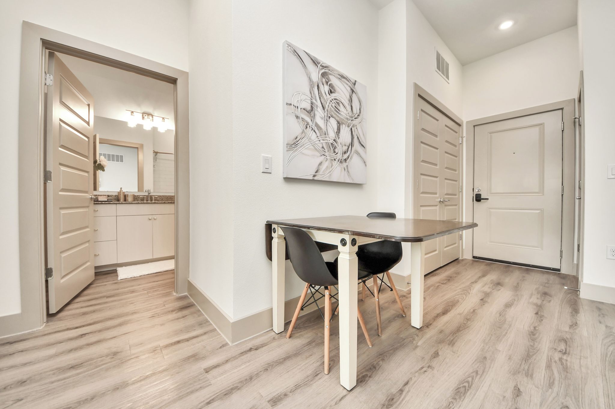 Un intérieur moderne comprenant un coin repas avec une table et des chaises, un couloir menant à une salle de bain et une porte dotée d’un miroir.
