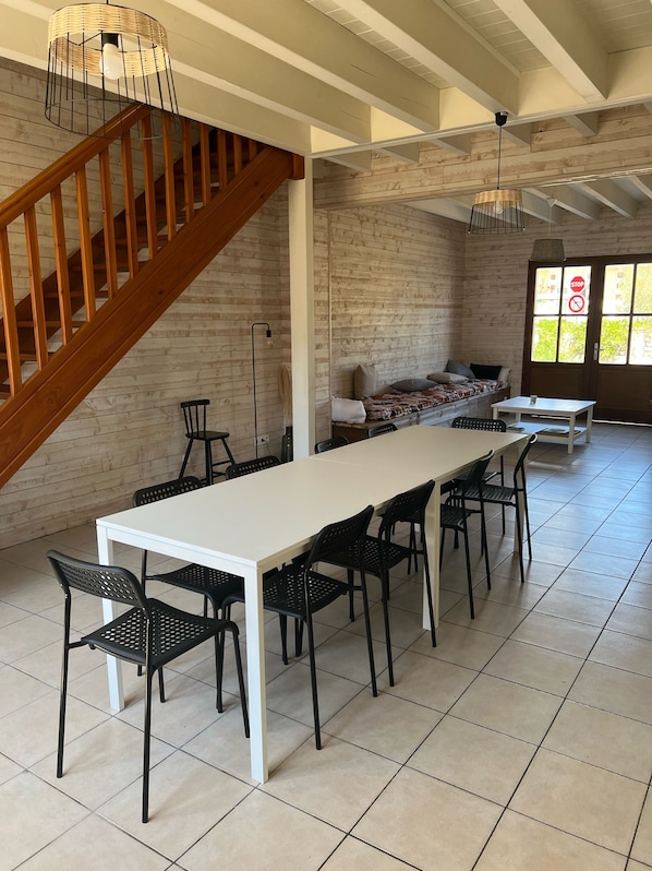 Une pièce spacieuse avec une longue table blanche, des chaises noires, un escalier en bois et un mur en briques.