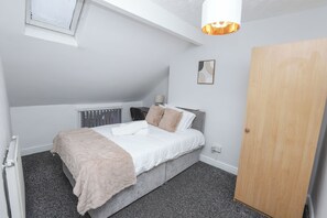 A bedroom with a bed, a bedside table, a lamp, a radiator, a wooden door, and a small wall art piece. - Prime Location 3 Bedroom Apartment Central (Middlesbrough)