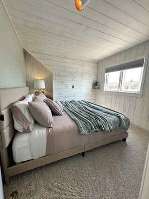 A bedroom with a bed, pillows, a bedside lamp, a window, and a plant. - Nice cozy 2-bedroom guest house in charming Marshfield. Short walk to the beach. (Marshfield)