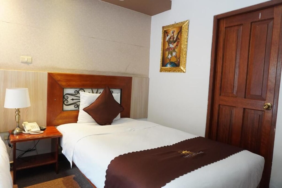 A hotel room with a bed, a bedside table with a lamp and phone, a door, and a wall-mounted picture.