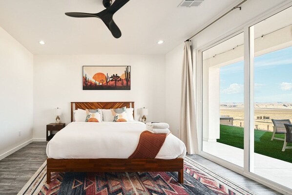 A hotel room with a large bed, a ceiling fan, a painting, and a view of a desert landscape through a large window. - The Red Canyon Retreat - Views, Hot Tub, Game Room (Page)