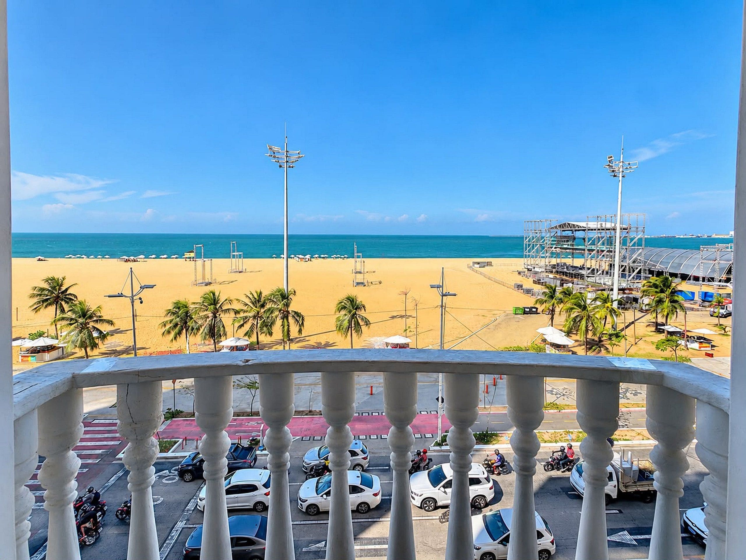 Een balkon met uitzicht op een strand, palmbomen en een parkeerplaats met auto's en motorfietsen.