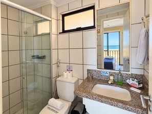 Een badkamer met een douchecabine van glas, een toilet, een wastafel met een granieten blad en een spiegel die een balkon met uitzicht op zee weerspiegelt.