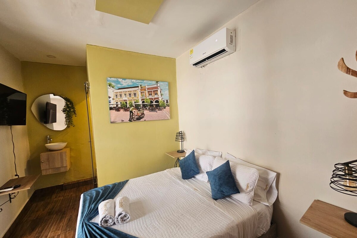 A hotel room with a bed, a bedside table, a mirror, a wall-mounted air conditioner, and a picture of a building.