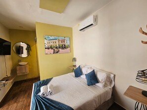 A hotel room with a bed, a bedside table, a mirror, a wall-mounted air conditioner, and a picture of a building. - Hotel Almabella (Cartagena de Indias)