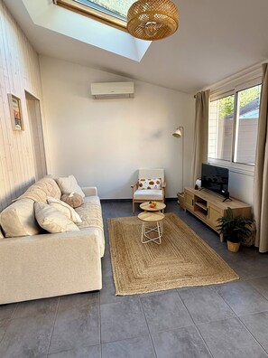 En moderne stue med sofa, lænestol, lille bord, fjernsyn og et vindue med gardiner.