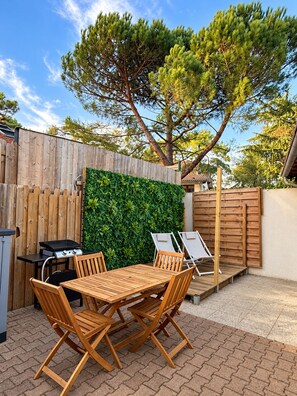 En udendørs terrasse med et træbord og stole, grill og en grøn væg.