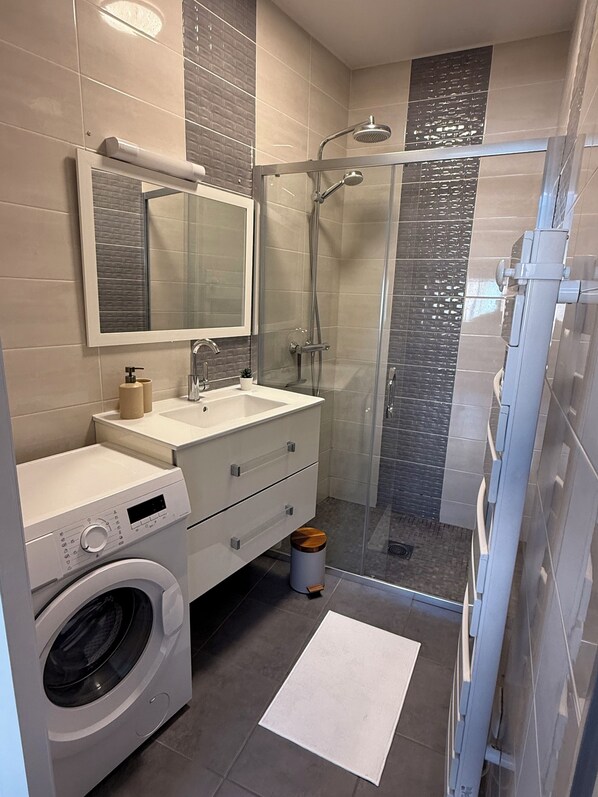 Ett modernt badrum med en inbyggd tvättmaskin, en duschkabin med glasväggar och en stor spegel ovanför handfatet.