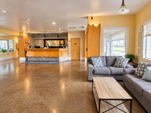 Una hall d'albergo con una reception, un divano grigio con cuscini decorati, un tavolino in legno e un'ampia finestra con persiane.
