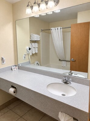 Un bagno d'albergo con uno specchio grande, un lavandino, un asciugacapelli e una tenda per la doccia.