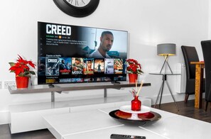 Uma sala de estar moderna com uma televisão de tela plana exibindo um filme, uma mesa de centro com um objeto decorativo e duas plantas em vasos.