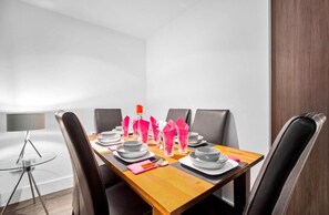 Mesa de jantar para quatro pessoas com design moderno, incluindo tampo de vidro, cadeiras escuras e guardanapos cor-de-rosa.