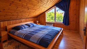Ein Schlafzimmer mit Holzboden, einem großen Bett mit blauer Bettwäsche, einem Nachttisch, einem Fenster mit Blick auf Bäume und einer Tür.