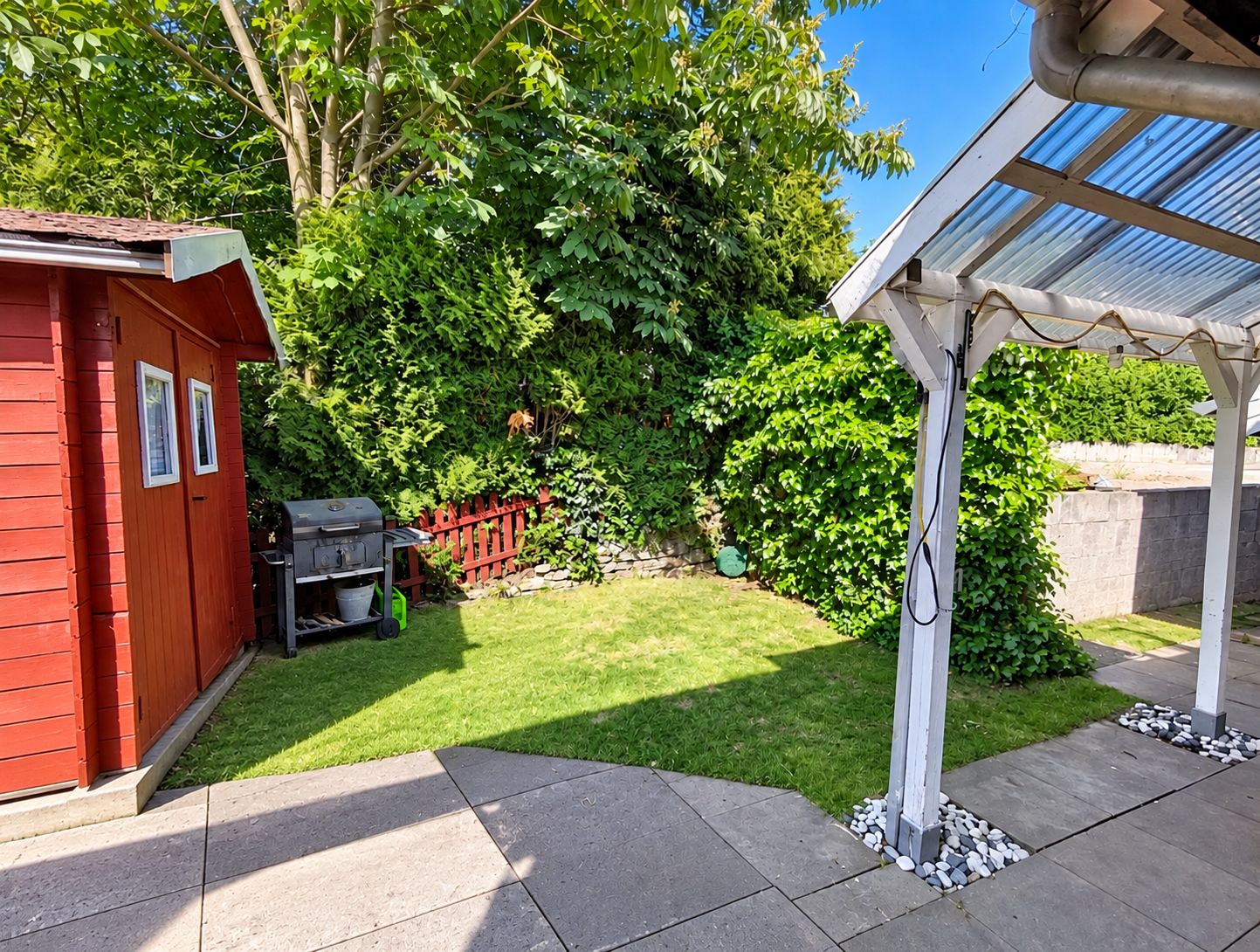 Ein Garten mit einem roten Gartenhaus, einem Grill, einer Pergola und einem gepflegten Rasen.