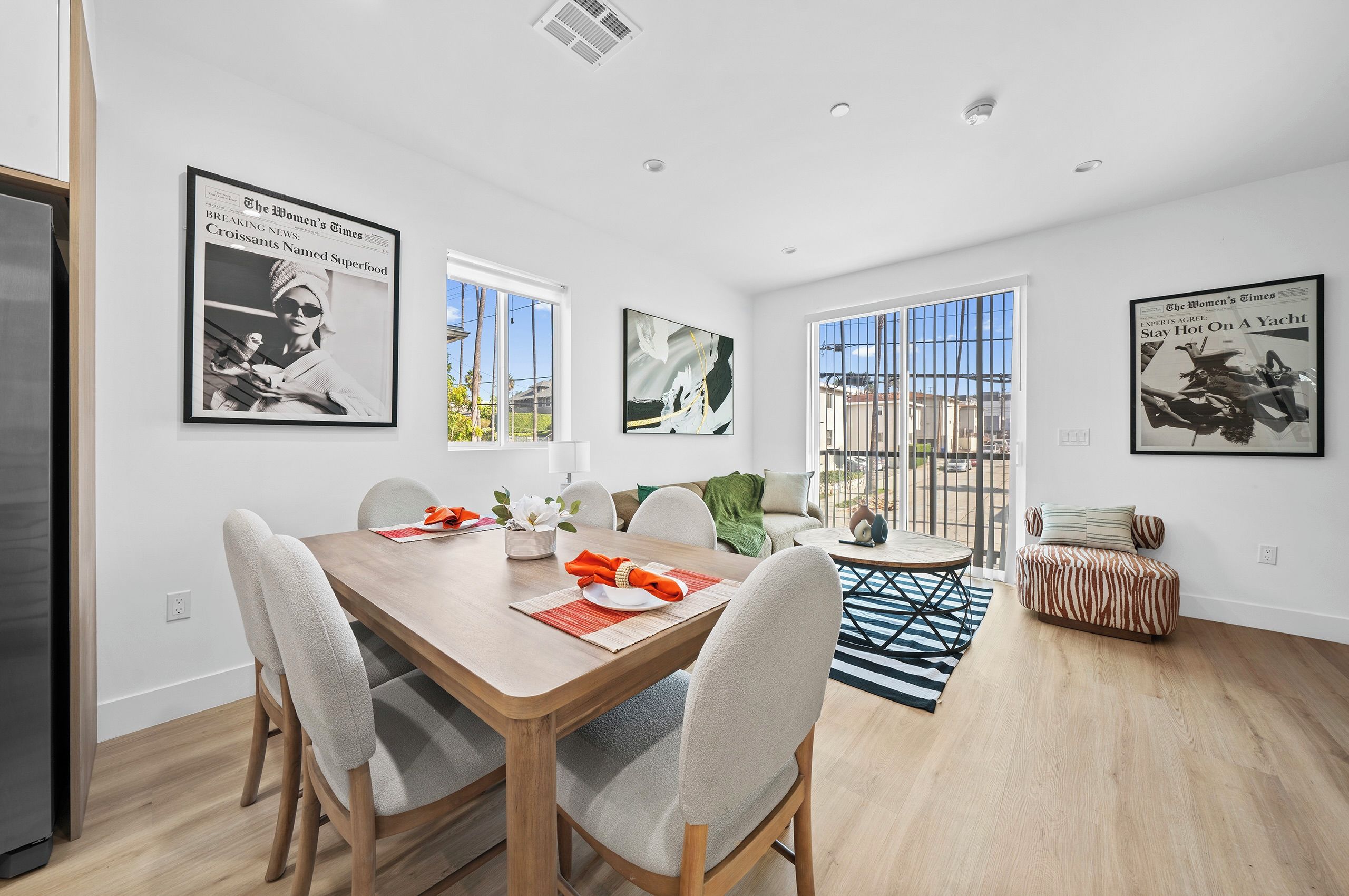 A modern dining area with a wooden table, white chairs, and a rug. There are framed pictures on the walls and a sofa in the background.