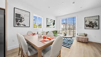 A modern dining area with a wooden table, white chairs, and a rug. There are framed pictures on the walls and a sofa in the background.