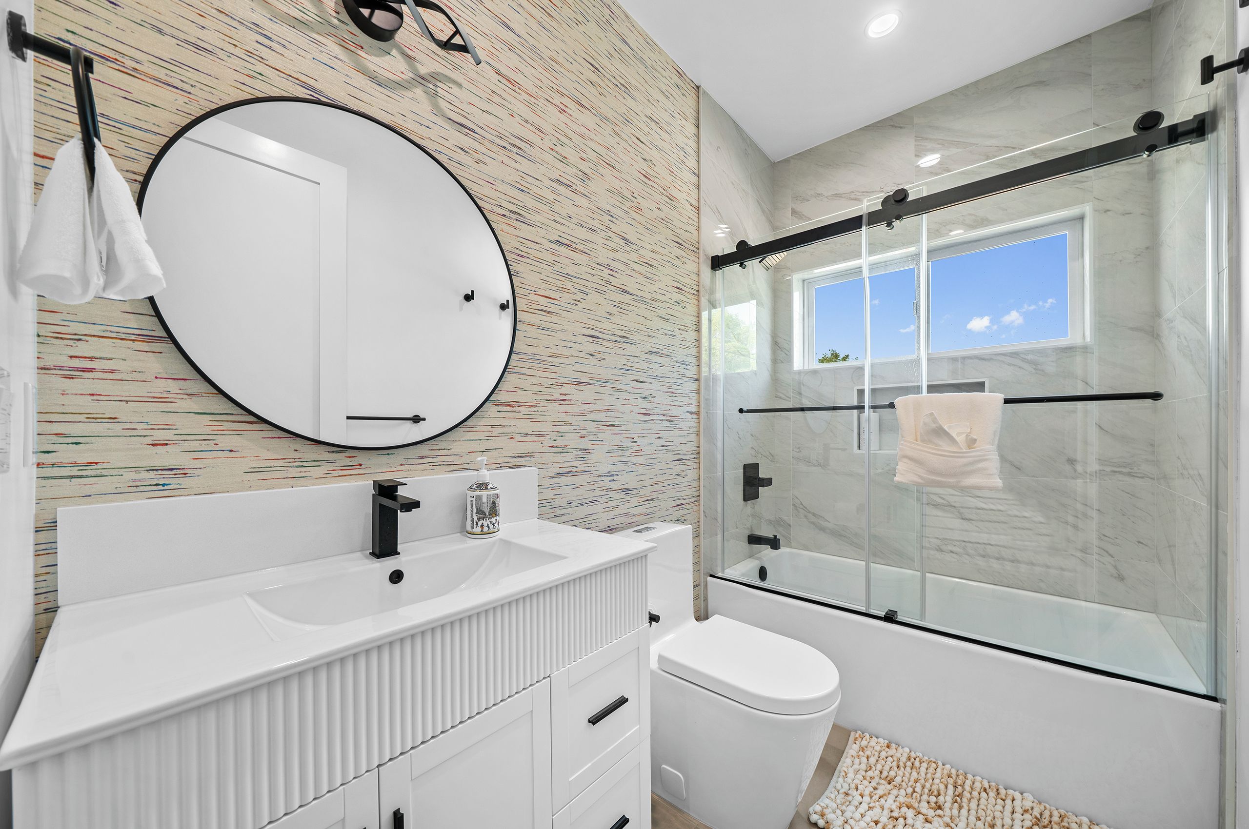 A modern bathroom with a large round mirror, a white sink, a glass shower enclosure, and a view of the sky through the shower window.