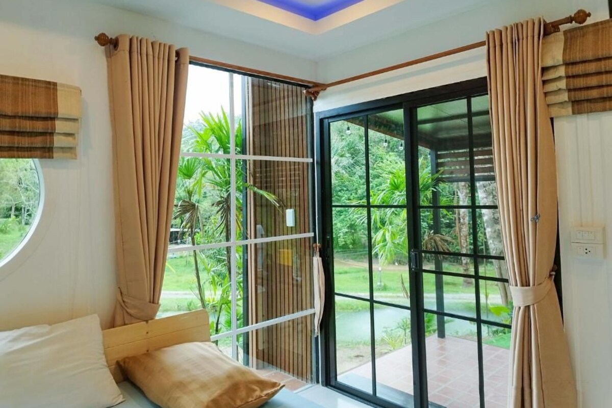 A room with a bed, a window, and a sliding glass door leading to an outdoor area with greenery.