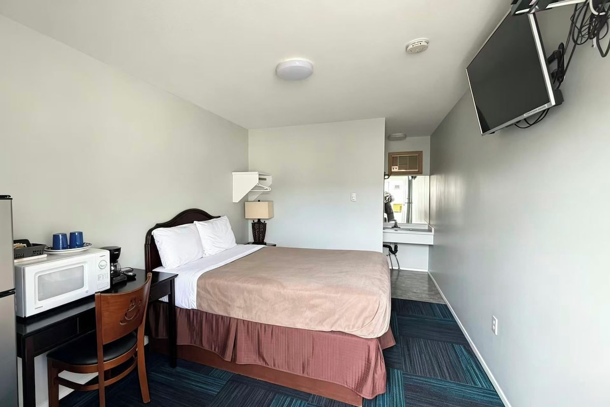 A hotel room with a bed, a desk with a microwave, a chair, a lamp, and a TV mounted on the wall.