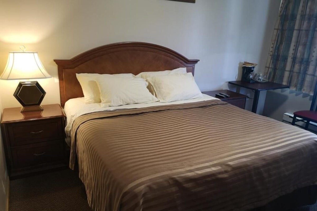 A hotel room with a bed, a nightstand with a lamp, a small table with a coffee maker, and a chair.