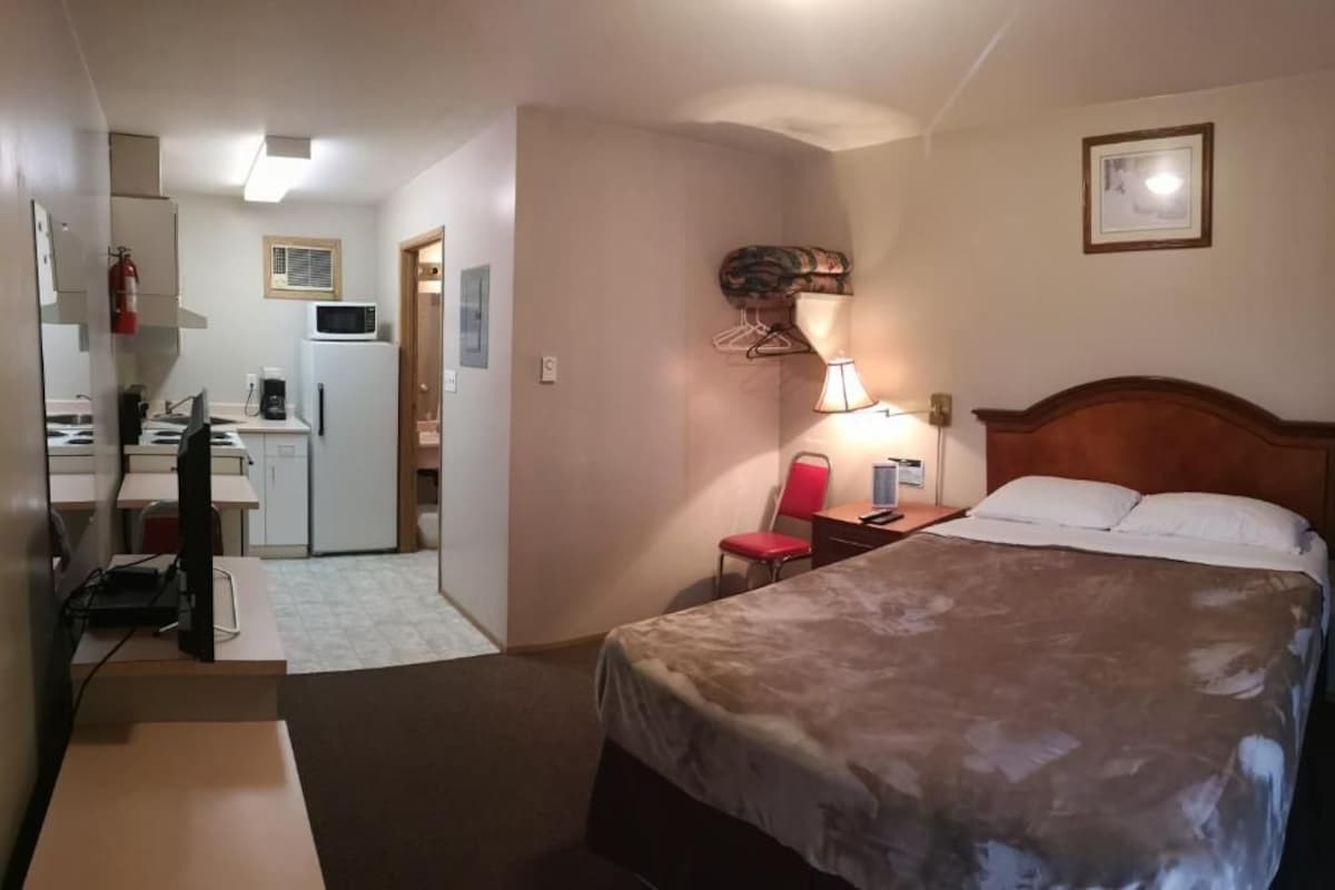 A hotel room with a bed, a desk, a chair, a lamp, a television, and a kitchenette.