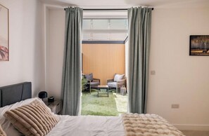 Une chambre à coucher comprenant un lit, une table de chevet, une lampe et une vue sur un espace extérieur aménagé, visible à travers une fenêtre avec des rideaux.