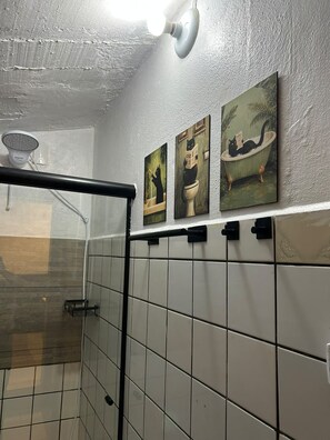 A bathroom with white tiled walls, a shower area, and three wall-mounted paintings. - New Listing (Jaraguá do Sul)