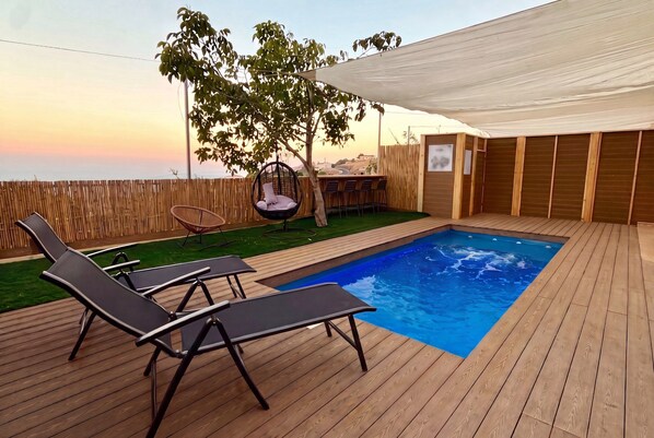 Une terrasse en bois avec une piscine, des chaises longues, un fauteuil suspendu et un espace couvert offrant une vue agréable.