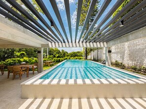 Ein rechteckiger Swimmingpool mit klar blauem Wasser, umgeben von einer Pergola mit horizontalen Balken und einer Terrasse mit Holzmöbeln.