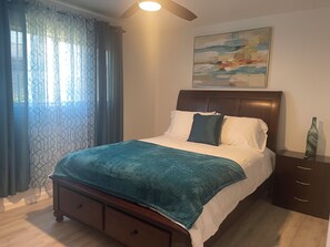 Une chambre à coucher avec un lit, une table de chevet, un tiroir, un tableau accroché au mur et une fenêtre avec des rideaux.