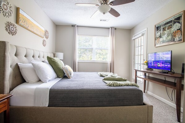 A bedroom with a bed, a ceiling fan, a television, and a window with blinds.