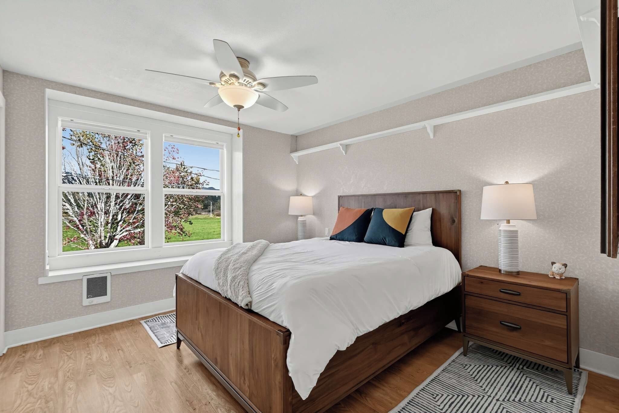 A bedroom with a bed, bedside tables, a ceiling fan, and a window with a view of trees.