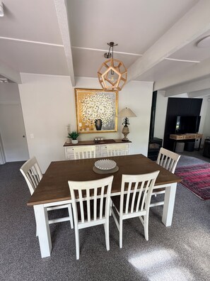 A dining area with a wooden table and white chairs, a chandelier, a framed artwork, and a television in the background. - Boutique BarnStay/Games room,Boat parking on beautiful private grounds (Taupō)