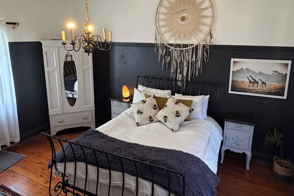 A bedroom with a bed, a chandelier, a dreamcatcher, a nightstand, and a rug.