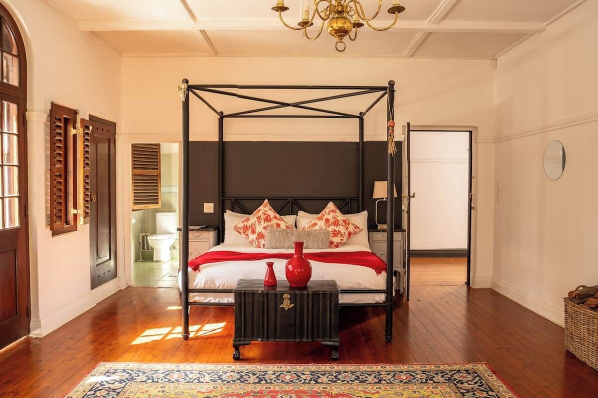 A four-poster bed with a dark headboard, a red vase on a side table, and a patterned rug on the floor.