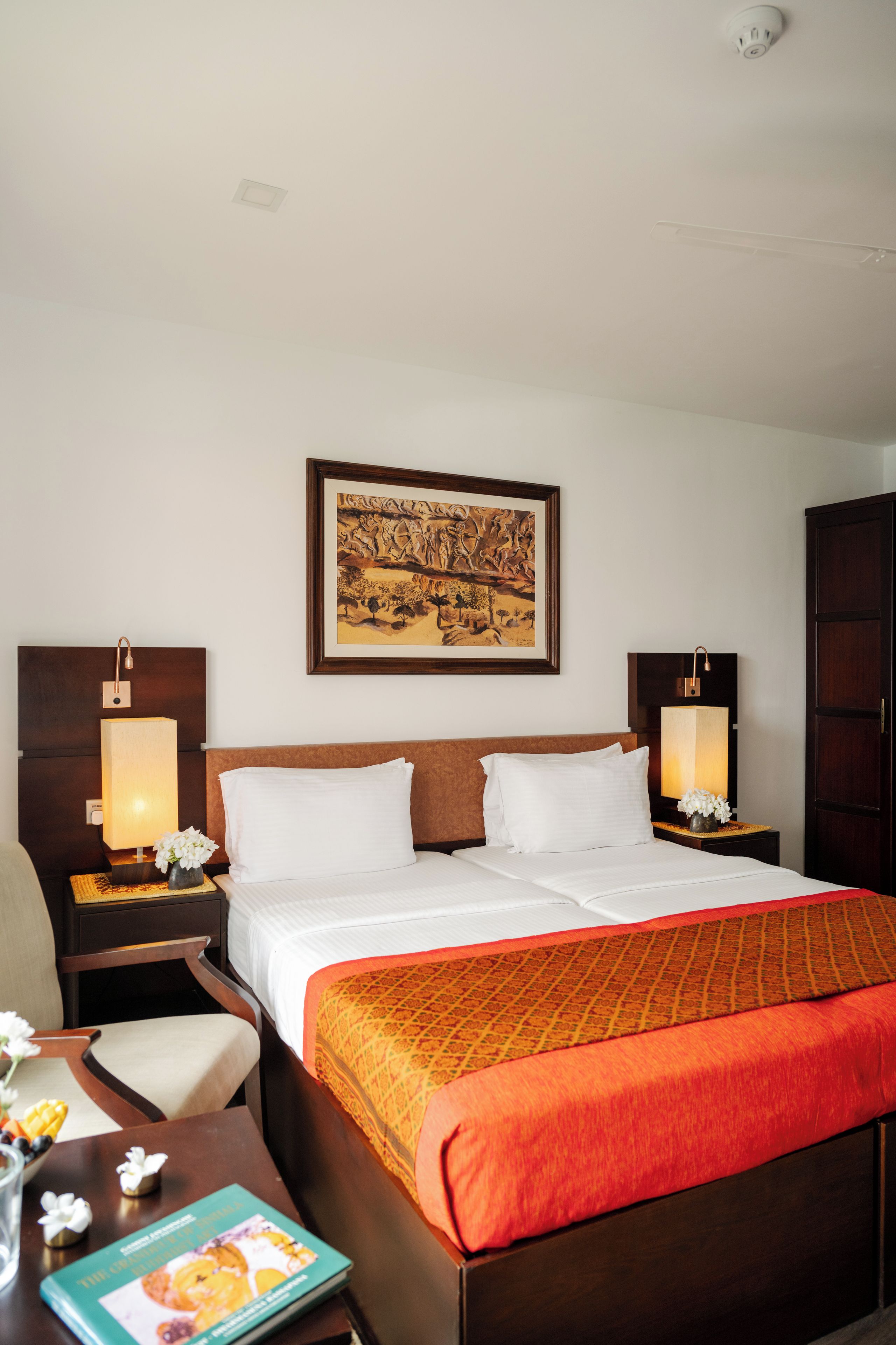 Una habitación de hotel con una cama grande, dos mesitas de noche con lámparas, una obra de arte enmarcada sobre la cama y un armario de madera.
