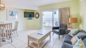 Una sala de estar con una mesa de centro de madera, un sofá, una silla y un televisor. Hay un balcón con vista al mar.