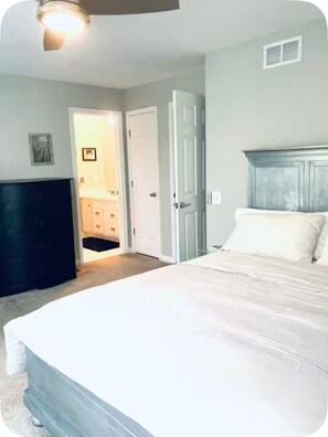 Een slaapkamer met een groot bed, een donker hoofdeinde, een witte deur en een gang naar een badkamer.