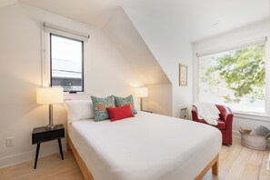 Een slaapkamer met een bed, twee bedlampen, een rode stoel, een raam en een aan de muur bevestigd schilderij.