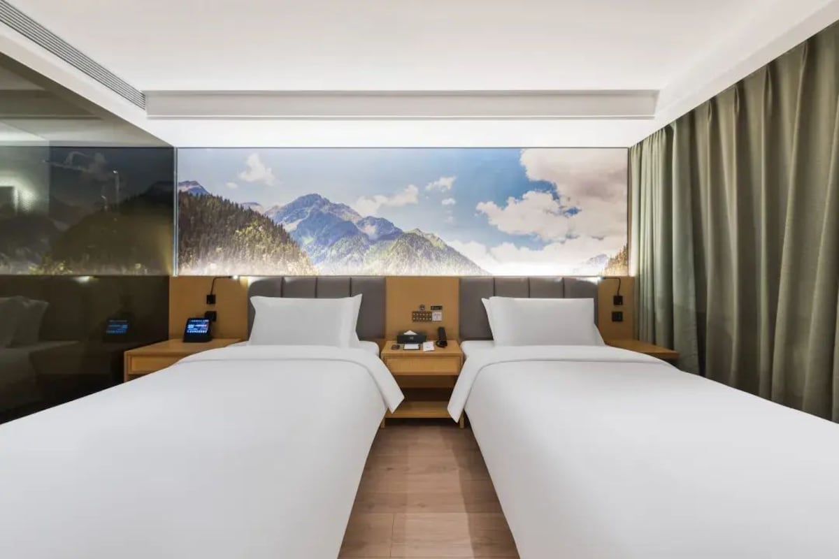 Habitación de hotel con dos camas, un ventanal con vista a la montaña, una mesita de noche con lámpara y un televisor montado en la pared.