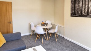 Een kleine kamer met een grijze bank, een witte tafel met stoelen, en een aan de muur hangend schilderij van een bos.