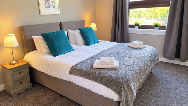 Een net opgemaakt bed met een grijs hoofdbord, wit beddengoed en teal kussens. Twee bedlampen staan op houten nachtkastjes, en een raam met gordijnen biedt uitzicht op groen.