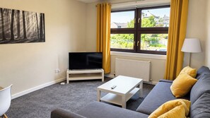 Een gemeubileerde woonkamer met een televisie, bank en een raam met gordijnen.