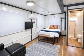 Kompakti hotellihuone, jossa on sänky, yöpöytä, penkki ja suihkutila.