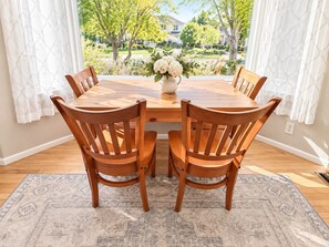 Un coin repas avec une table et des chaises en bois, un vase rempli de fleurs et une vue sur un quartier résidentiel, visible à travers de grandes fenêtres.